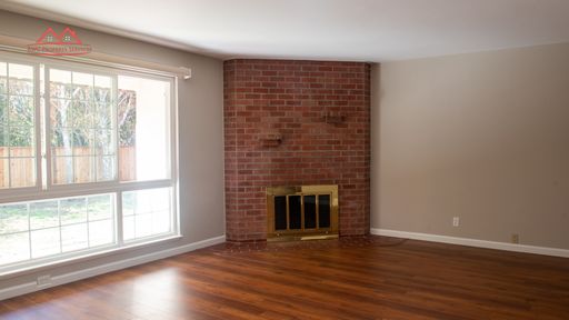 Living room with brick corner fireplace, hardwood flooring, and large windows